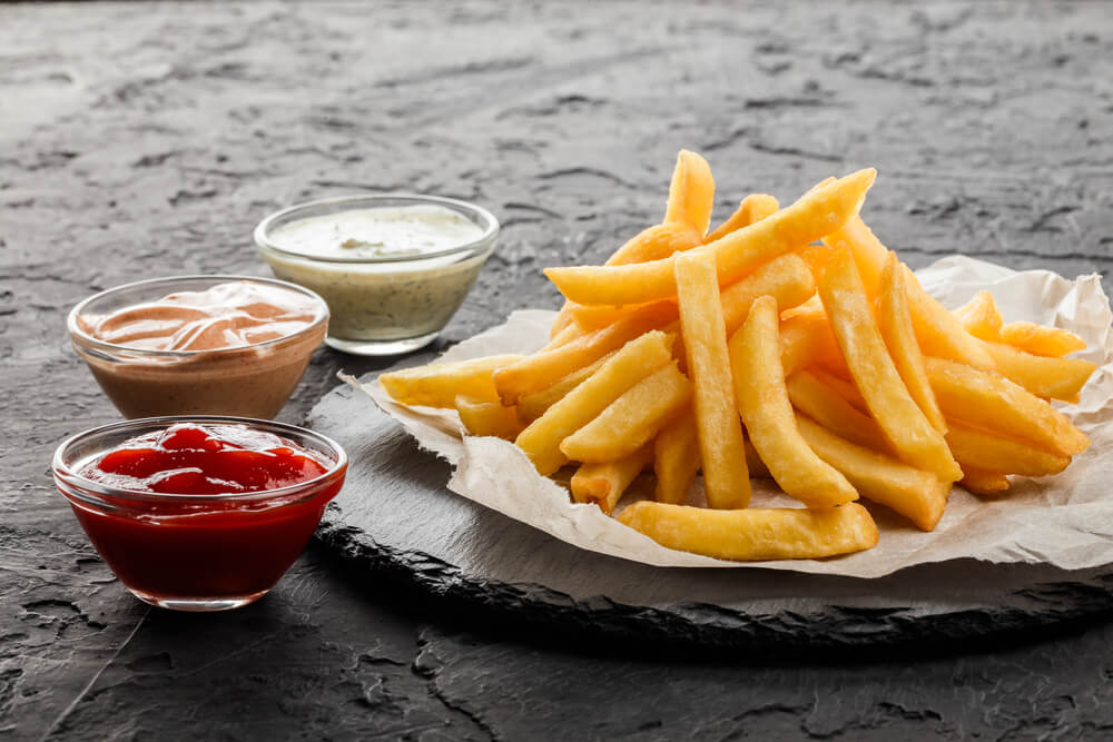 Tasty French Fries Potatoes on Paper With Sauces Over Black Stone Background