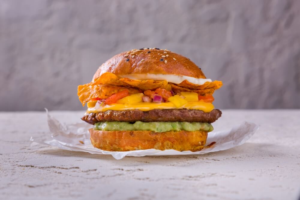 Hamburger With Cutlet, Mango, Salmon, Yellow, Sauce, White Sauce, Green Sauce, Onions and Chips That Stands on White Paper on a Rough Concrete Table
