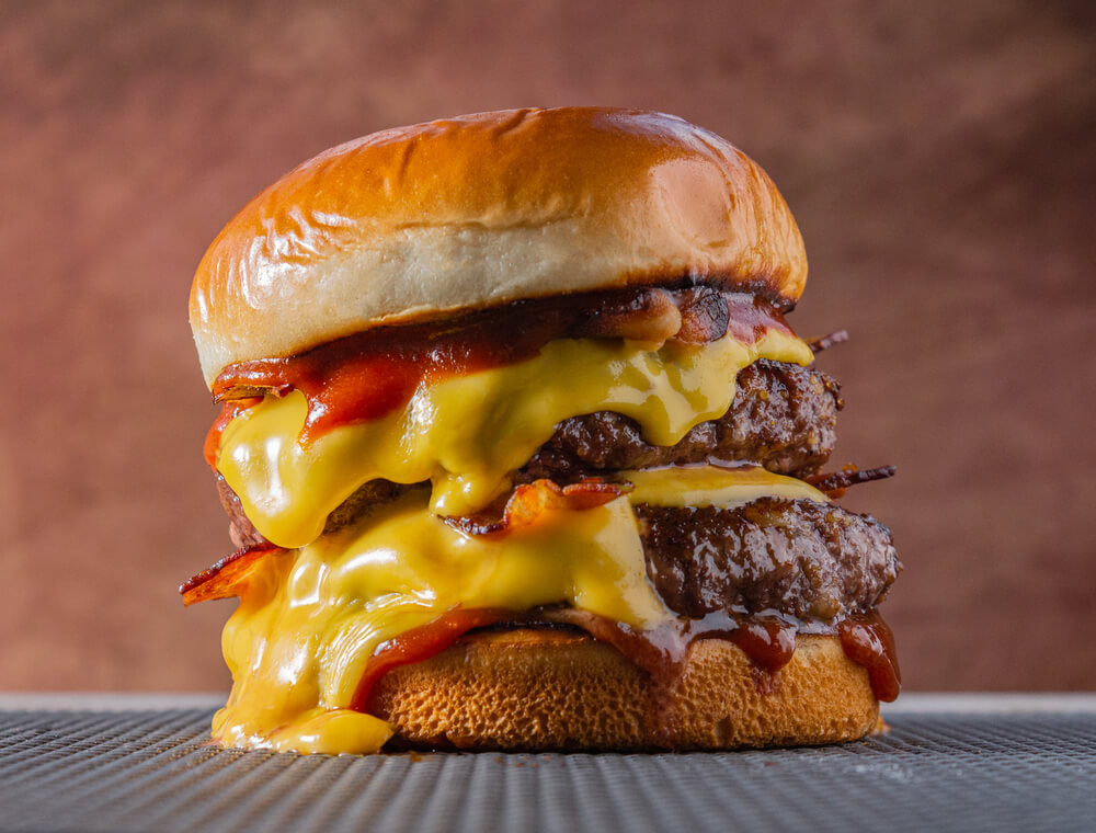 Double Meet Burger With Melting Cheddar Cheese and Bacon Centralized on a Gray Surface and Brown Background